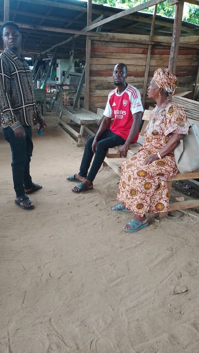 Three people are present. A man stands on the left wearing a patterned shirt and dark pants. Another man sits in the middle wearing a red and white soccer jersey and dark pants. An elderly woman sits on the right wearing a patterned dress and head wrap. They are sitting or standing near a wooden structure with other benches. The ground is sandy. The location is Asubi, Nigeria.