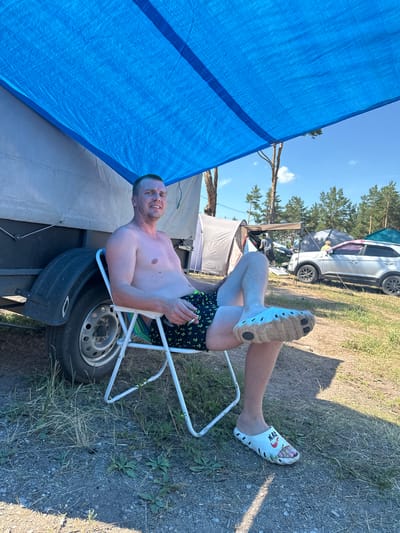 A man is sitting on a folding chair under a blue tarp in a campsite. He is shirtless and wearing black swim trunks with an anchor pattern. His legs are crossed, and his feet are covered in white clogs. To his left is a trailer with a black fender. In the background, there are tents, trees, and a silver car. The ground is covered in dirt and grass. The sky is blue with some clouds.
