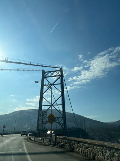 The image displays the eastern approach to the Bear Mountain Bridge in the Town of Cortlandt, New York, United States of America. A prominent metal lattice truss bridge tower stands centrally, supporting suspension cables. In the foreground, a curved two-lane asphalt road, partially covered with what appears to be frost or ice, leads towards the bridge, bordered by a stone wall and a metal guardrail on the right. A white SUV is visible on the road ahead, moving towards the bridge. A red octagonal STOP sign is positioned on the left side of the road. Multiple rectangular white road signs beneath the bridge tower indicate "EAST/WEST ROUTE 6" and "EAST/WEST ROUTE 202." Below these, a yellow diamond-shaped sign warns of an "ICY PAVEMENT ZONE." The sky is clear blue with scattered white cumulus clouds and a single prominent airplane contrail diagonally traversing the upper left quadrant. A bright sun flare emanates from the upper left. Snow-dusted mountains form the backdrop, with the western span of the bridge visible against them.