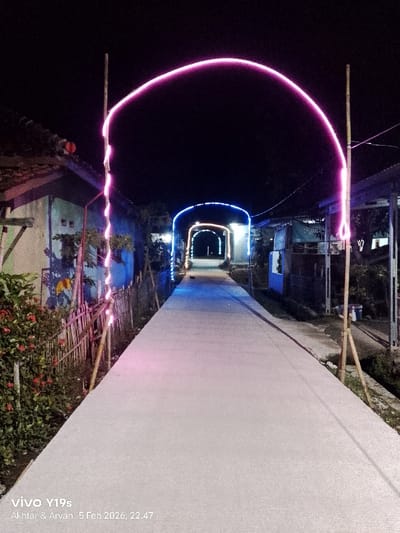 A pathway illuminated by a series of lighted arches stretches into the night. The initial arch glows with a vibrant pink, succeeded by arches with blue and yellow lights. Dwellings flank both sides of the path, appearing as silhouettes in the darkness. The pathway, likely concrete, is a dominant feature in the foreground. The date 5 Feb 2026 and other details are visible at the bottom.