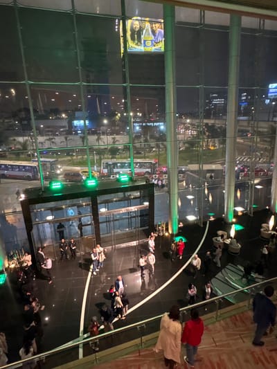 The image depicts an aerial view of a plaza or public space in San Borja, Peru, at night. The perspective is from a high vantage point, possibly within a building looking down. Numerous people are scattered across the plaza, walking or standing in groups. The plaza has a shiny, reflective surface that reflects the ambient light and the figures of people. Glass panels and green architectural supports are visible in the foreground, providing a frame for the view of the city and the plaza. There is a billboard with a picture on the left side of the image. City lights and buildings can be seen in the background, indicating the urban location.