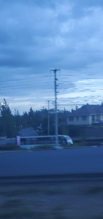 This image captures a semi-urban landscape in Kinanie ward, Kenya, under dim, overcast conditions with a predominant blue-grey hue. The scene appears to be documented from a moving vehicle, indicated by motion blur in the foreground and across central elements.

The upper approximately 40% of the frame shows a sky filled with dark grey-blue clouds. A prominent, slender electrical pylon with multiple cross-arms and numerous power lines extends vertically near the center of the image, spanning from the mid-ground almost to the top.

In the mid-ground, a dark grey-blue paved road traverses horizontally across the frame. On the far side of this road, a light-colored, possibly white or light grey, passenger bus or coach is visible, exhibiting motion blur and partially obscured by the electrical pylon. Behind the road and the pylon, a dense line of dark green, coniferous trees forms a significant portion of the background. To the right, behind the tree line, a two-story building with a dark, sloped roof and light-colored walls is discernable. Other smaller utility poles and lines are present amidst the trees.

The foreground, occupying the bottom approximately 15% of the frame, consists of indistinct dark ground, also affected by motion blur. No individuals are visible. The overall lighting suggests either twilight or heavily clouded daytime conditions.