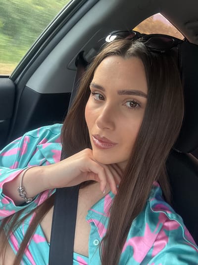 A medium close-up selfie shows a woman with long brown hair sitting in the passenger seat of a car, looking at the camera. She is resting her chin on her hand, which is adorned with a silver bracelet with a charm. She is wearing a teal and pink star-patterned button-down shirt and a black seatbelt. Sunglasses are perched on top of her head. The background is blurred, showing greenery through the car window and the car's interior. The setting is not specified as Draganovtsi, Bulgaria.