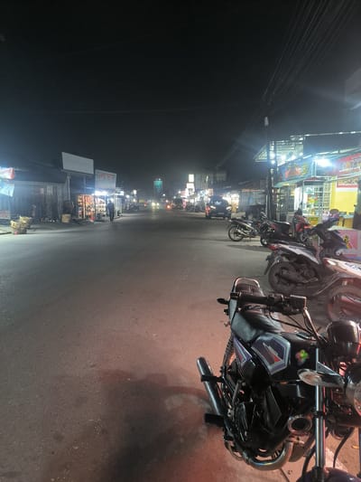 The image depicts a street scene at night in Jamurujung, Indonesia. The street is lined with small businesses, many of which are illuminated, suggesting commercial activity. In the foreground, a black motorcycle with purple and silver accents is parked, its chrome details reflecting the surrounding lights. Several other motorcycles are visible parked along the roadside and on the street further down.
A lone figure is walking away from the camera on the right side of the street, and other figures appear to be present in the doorways of the shops. Some of the shops have awnings or roofs extending over the sidewalk. Baskets, possibly for goods, are stacked near one of the shops. The street itself is asphalt and appears wet in places, reflecting the overhead lights.
The sky is dark, indicating it is nighttime. The lighting from the shops and streetlights creates a warm glow on the scene. Visible text includes "RAHMAT GESTRO" on a sign above one of the shops. Other signs are present but are too blurry to read. The overall atmosphere suggests a lively evening in a commercial area. The weather appears clear.