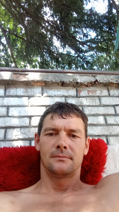 A man is shown in a close-up selfie, taken from chest up, outdoors. He is shirtless and appears to be resting against a red, fuzzy object, possibly a cushion or blanket. Behind him is a weathered brick wall, and above that, branches of a tree obscure the sky. The setting suggests a casual, possibly backyard, environment. The man has short, dark hair, a light complexion, and is looking directly at the camera with a neutral expression. The image's composition and lack of context do not provide information about the specific city of Altai, Kazakhstan, or any distinguishing urban features.