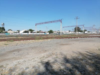 The image shows a view of a train track area in the city of Juma, Uzbekistan. In the foreground, there is a dirt and gravel path with scattered stones and dry grass. Beyond the path are multiple parallel train tracks, with some vegetation growing between them. A blue metal fence runs along the edge of the tracks. In the background, there are several buildings, including houses with white and brown roofs, and some trees. Overhead, there are numerous power lines and supporting structures, including tall red metal gantries for the overhead power supply to electric trains, and concrete poles with crossbeams supporting wires. A water tower is visible in the distance on the left. The sky is clear and blue. The scene appears to be an outdoor, daylight shot, likely taken during the day. There are no people visible in the image.