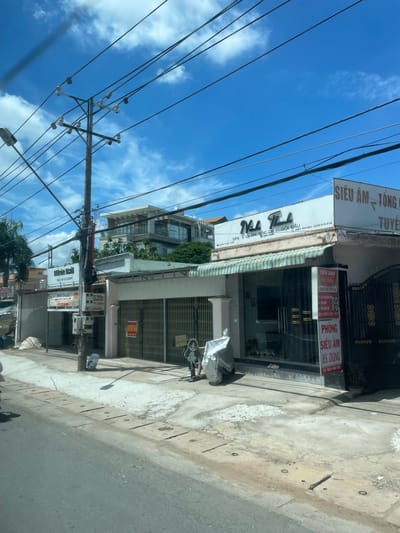 The image shows a street scene in Phường An Tịnh, Vietnam, under a bright blue sky with scattered clouds. Utility poles with numerous wires are prominent in the foreground and midground, crisscrossing the sky. Along the street, there are several businesses. To the left, a building with a sign reading "Minie Nail" is visible, with smaller signs below advertising prices. Further to the right, a building with a large glass storefront has signage that includes "Nha Thanh SPA" and "Chăm Sóc Da" (Skin Care). Adjacent to this is another building with a sign in Vietnamese that appears to be for a "Phòng Siêu Âm" (Ultrasound Room). A gate made of gold-colored metal is to the far right. The street in front of the businesses is paved with concrete and asphalt, with some areas showing wear and tear. A covered object resembling a motorcycle or scooter is parked on the sidewalk. Various trash receptacles and smaller signs are also present. A modern multi-story building with balconies and glass windows is visible behind the street-level businesses.