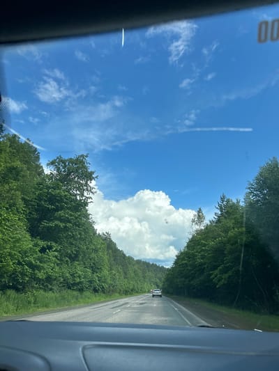 This is a photo of a road in the city of Usman-Gali, Russia, on a cloudy day. The road is paved and has two lanes of traffic. There are trees on either side of the road. The sky is blue with some white clouds. The photo appears to have been taken from inside a car. There are other cars on the road, but no people are visible.