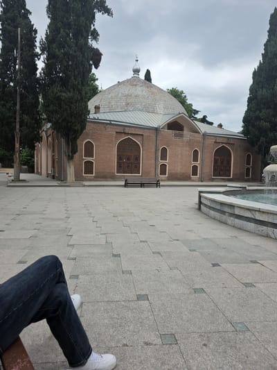 The image shows a brick building with a domed roof and arched windows, possibly a mosque or mausoleum, in Ganja, Azerbaijan. The building has intricate wooden latticework on its windows. To the right of the building, a large tiled courtyard leads to a fountain with flowing water. Green trees are visible behind and to the left of the building. In the foreground, a person is sitting on a bench, their legs crossed, wearing dark jeans and white sneakers. The overall scene suggests a calm and serene atmosphere. The sky is overcast, indicating a cloudy or potentially rainy day. The time of day is difficult to determine due to the lighting conditions. There is no visible text in the image.