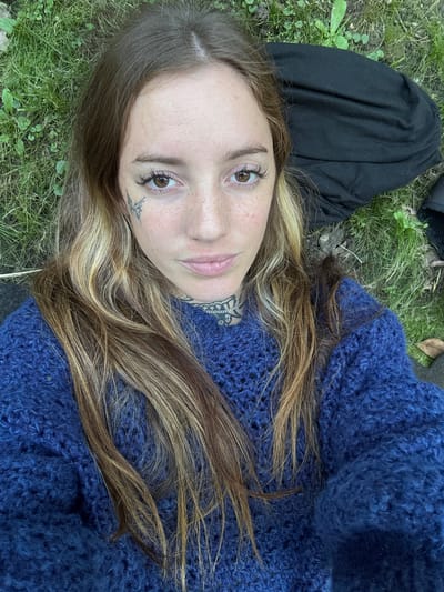 A woman is lying on her back on grass. She is wearing a blue sweater. She has brown and blonde hair, brown eyes, and a tattoo below her left eye and on her neck. Part of what appears to be black clothing is visible to the left. The location is Villa General Belgrano, Argentina.