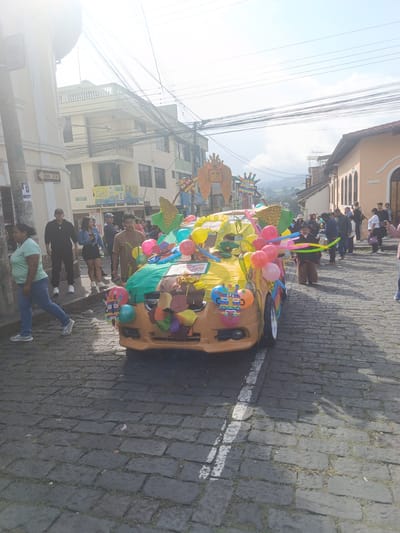 In Amaguaña, Ecuador, a daytime street scene depicts a festive procession. The central subject is a light brown/tan sedan, extensively decorated with colorful balloons (pink, yellow, blue), green and yellow fabrics, cut-out shapes resembling leaves and fruits, and mask-like figures affixed to the front, sides, and roof. A large golden, corn cob-like structure and an ornate, winged sun-mask adorn the roof.
Numerous individuals populate the street and sidewalks. In the left foreground, a woman in a light green t-shirt and blue jeans walks from left to right. Mid-left, a man in a dark tracksuit stands on the sidewalk, while a woman in a denim jacket examines a mobile phone. On the right side of the decorated vehicle, a young boy in a dark hat and brown fur-like leggings walks alongside, holding elongated, colorful balloons. Spectators and participants, comprising adults and children, line both sides of the cobblestone street further in the background.
The environment features dark, irregular cobblestone pavement with sparse grass, and a faded white lane marking on the right. Buildings on the left include multi-story structures with light-colored facades, one striped vertically in yellow and white, displaying a "UNA VIA" (One Way) sign and another with a "CENTRO DE EDUCACION MI PRIMER ALEGRIA" sign. Buildings on the right exhibit peach-colored facades with arched windows and terra cotta tiled roofs. A dense network of black electrical wires is strung overhead. Rolling hills or mountains are visible in the distant background under a partly cloudy sky, with a sun flare present in the upper left. The decorated car is in motion or stationary as part of an event, with people observing.
