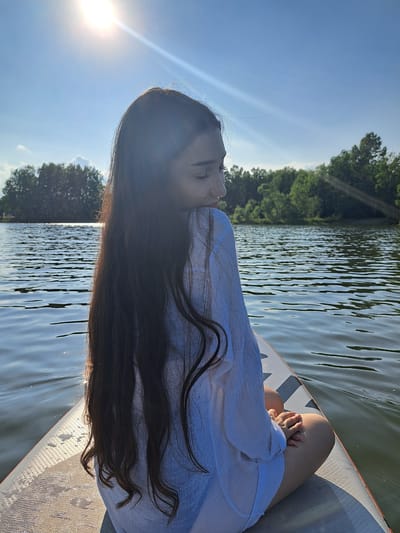 A young woman with long, dark hair sits cross-legged on a paddleboard on a calm body of water. She is facing away from the camera, looking to her right, with her head tilted down. The sun is bright in the upper left corner of the image, casting a lens flare. The water is rippled, reflecting the blue sky and surrounding trees. The paddleboard is light-colored with a grey patterned grip. The woman wears a loose-fitting white shirt. The background features a line of lush green trees. The scene appears to be set in a natural, outdoor environment, possibly a lake or river. The city of Раздольное, Russia is not visible in the image.