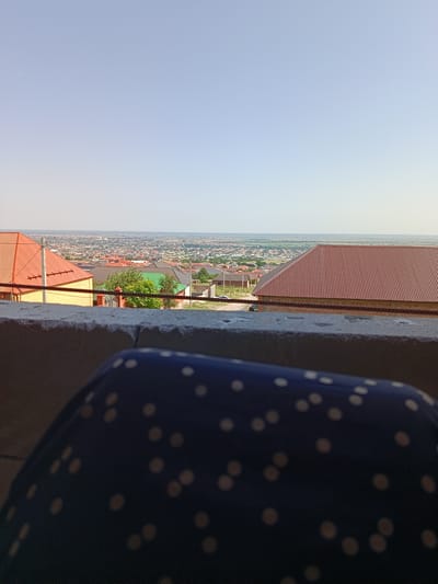 A high-angle, close-up shot focuses on a dark blue fabric with a pattern of light-colored dots. In the background, a panoramic view of a city or town unfolds under a clear, pale blue sky. The cityscape features numerous houses with red and brown roofs, interspersed with green trees and a distant horizon. The foreground is partially occupied by a concrete ledge or wall, with a twisted rope or wire draped across it. The overall impression is one of an elevated vantage point overlooking a residential area on a bright, clear day. The image appears to be taken in Novyy Benoy, Russia, given the architectural style and landscape.