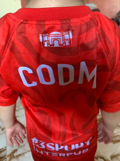 The image displays the rear view of an individual, likely a child or young person, standing indoors in Sidi Slimane Moul Al Kifane, Morocco. The subject is wearing a red short-sleeved t-shirt with a subtle, darker red wavy or striped pattern integrated into the fabric. Near the upper back of the shirt, a white stylized architectural graphic depicting a building with multiple domes and arches is visible. Directly below this graphic, the acronym "CODM" is prominently displayed in large, white capital letters. Towards the lower back, partial white text reads "S3SPORT" or "S3SPURT" above "INTERPUB". The person's arms are extended slightly outward, with both hands visible on either side of the lower torso. The floor beneath the subject consists of light-colored tiles, possibly with a marble-like pattern. The background is blurred, indicating an indoor environment.