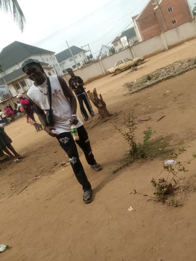The image depicts an outdoor scene in Umuokpu, Nigeria, characterized by an unpaved, reddish-brown dirt ground under an overcast sky.
In the foreground, a dark-complexioned male, estimated to be in his twenties, is positioned centrally, smiling and looking directly at the camera. He wears a black beret, a white sleeveless tank top, black jeans with white pattern details, and grey open-toed sandals. A black cross-body bag strap is visible over his left shoulder, and he wears a beaded necklace and a watch on his left wrist. His right hand holds a light-green plastic bottle with a white straw.
To the immediate left of the main subject, partially obscured, is a group of individuals, some wearing pink attire, appearing to be gathered or seated on the ground. Further into the midground, to the right of the main subject, another dark-complexioned male stands, wearing a black t-shirt and black pants with white side stripes, holding a dark object in his right hand.
The ground consists of dry, dusty dirt with scattered small green shrubs and two prominent dark brown tree stumps. A discarded clear plastic bottle lies on the ground in the lower left.
In the midground, a low, light-colored concrete wall extends horizontally. Behind this wall, an older model, light-yellow sedan, possibly a Peugeot, is parked on a dirt surface. Beyond the wall and car, various buildings are visible. These include a multi-story building with a dark, pitched roof and white walls to the left, and a partially constructed or uncompleted brick building with light-colored walls to the right. Overhead electrical wires crisscross the sky. A partially visible white banner with blue text is affixed to a building in the background, and a sign displaying "HAIR AND..." can be discerned on another background structure.