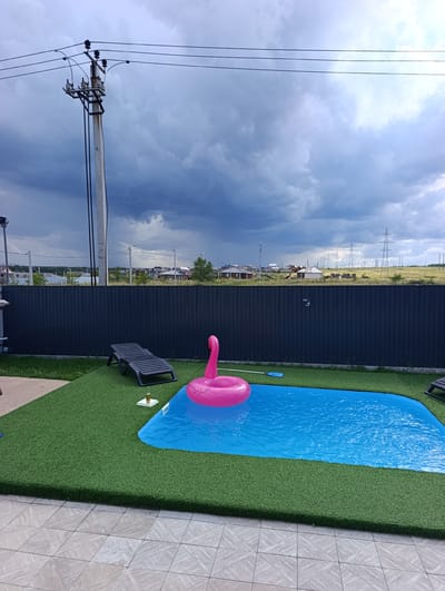 A backyard scene depicts a blue swimming pool with a pink flamingo inflatable ring floating in it. The pool is surrounded by artificial green grass. A dark-colored lounge chair is positioned on the grass near the pool. In the background, a dark grey fence runs horizontally across the frame, separating the yard from a residential area with houses and power lines. The sky is overcast with dark, dramatic clouds, suggesting an impending storm. The overall environment appears to be a suburban or rural residential area in Ленина, Russia, characterized by the presence of residential buildings and utility infrastructure.