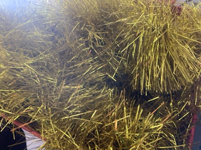 The image is a close-up, overhead shot of a large pile of gold tinsel. The tinsel is highly reflective and appears to be made of thin, metallic strands. The lighting is bright, with a strong glare in the upper left corner of the image, suggesting it's artificial light or sunlight. The tinsel is densely packed and has a somewhat chaotic, tangled appearance, with strands pointing in various directions. No people, specific objects, or environmental context beyond the tinsel itself are visible. The location in Fajo 2 Division Range, Nigeria, is not discernible from the visual information presented in the image.