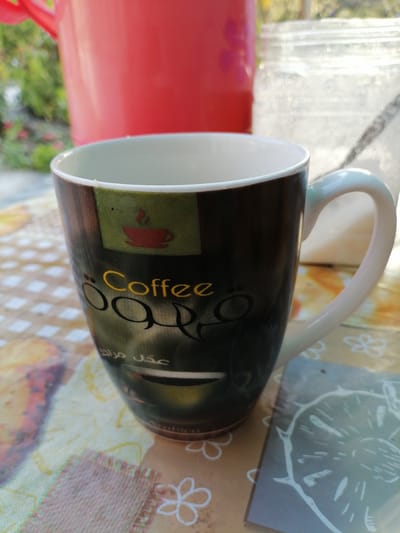 A close-up, eye-level shot captures a coffee mug with a brown and green design featuring Arabic script and a depiction of a steaming coffee cup. The mug's handle is white. It rests on a table covered with a patterned cloth. In the background, a large, out-of-focus red object, possibly another container, is visible, along with a blurred white object that could be a jar or bottle. The scene appears to be outdoors, with hints of green foliage and natural light. The overall impression is one of casual outdoor dining or relaxation. There is no information that allows for the determination of the location in Gumbaz, Uzbekistan.