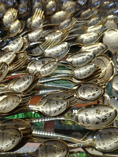 The image shows a collection of commemorative, silver-colored spoons and forks, possibly displayed for sale in Verkh-Sosnova, Russia. Each utensil is decorated with gold trim and gold floral accents. The spoons and forks each display a name in Cyrillic. Visible names include Anastasia, Vera, Varvara, Valentina, Valeriya, Vasilisa, Veronika, and more.