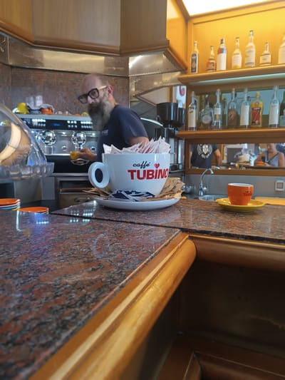 The image shows the interior of a bar, likely in Casalmaggiore, Italy, with a focus on the counter area. A man with a beard and glasses, wearing a dark t-shirt, is partially visible behind the counter, operating a coffee machine. In the foreground, a large white mug with the "CAFFÈ TUBINO" logo and a heart symbol is prominent, filled with sugar packets and stirrers. Beside it, a small orange espresso cup sits on a yellow saucer, also bearing the "TUBINO" logo. The counter is made of speckled red and black granite. Shelves behind the counter are stocked with various bottles of alcohol. Another person is visible in the background, reflected in a mirror, and the general ambiance suggests a busy cafe or bar environment.
