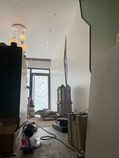 The image depicts an interior scene under renovation. In the foreground, to the left, is a dark, tall cabinet or piece of furniture. In the center, the floor is cluttered with construction materials including cardboard boxes, a bag of screws, and electrical cords. A large, tan storage container is visible on the right side of the frame, partially obscuring a white panel. Further back, a glass door covered with a white tarp leads to an outdoor area. Several ladders, including a multi-position ladder, are set up in the middle ground. A distinctive modern chandelier with multiple spherical lights hangs from the ceiling in the upper left quadrant. The walls are mostly white, with one section painted a muted green. The overall impression is one of an unfinished space. The location in Meaux, France, is not discernible from the visual information provided.