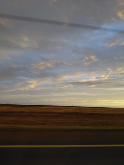 This image depicts a wide, open landscape under a cloudy sky, characteristic of the Prirechny Rural District in Kazakhstan. The foreground is dominated by a dark asphalt road with a distinct yellow line marking its edge. Beyond the road, a field of dry, brownish-yellow grass stretches towards the horizon. In the distance, a faint line of what appear to be wind turbines or utility poles can be seen against the muted tones of the landscape. The sky is a mix of gray and pale blue clouds, with soft, warm hues of yellow and orange peeking through, suggesting either sunrise or sunset. There are no people or active subjects visible in the frame. The overall impression is one of a rural, possibly somewhat arid, environment.