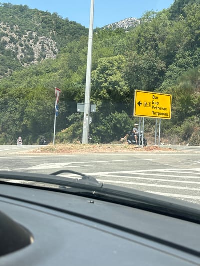 The image shows a road intersection in a mountainous, green environment, likely in or near Stupovi, Montenegro, based on the directional sign. The sign, which is yellow with black text, indicates directions to "Bar", "Бар", and "Petrovac" with the route marker "M-1". A man is seated on a scooter to the left of the sign. In the foreground, a car's windshield and dashboard are visible, suggesting the photo was taken from inside a vehicle. Other road signs, including a yield sign and a circular speed limit sign showing "40", are also present. The background consists of a steep, tree-covered hillside and a bright blue sky.