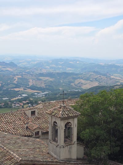 The image shows a view from a high vantage point in the city of San Marino, San Marino. In the foreground, there are several terracotta-tiled rooftops, typical of the region's architecture. A small bell tower with arched openings and a cross on top is prominent in the mid-foreground. To the right, a large green tree partially obscures the view. Beyond the rooftops, a vast, rolling landscape unfolds, characterized by green hills, cultivated fields in varying shades of brown and green, and distant mountains shrouded in a light haze. The sky is mostly clear with scattered white clouds. In the distance, a small town or village can be seen nestled in the valley. The overall impression is one of a serene, scenic vista of the San Marino countryside.