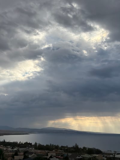 The image depicts a dramatic sky over a landscape in Kaji-Say, Kyrgyzstan. The sky is filled with a mix of dark, heavy clouds and patches of bright sunlight breaking through. These sunbeams create a strong contrast, illuminating the sky with a golden hue in some areas, while others remain dark and moody.

The scene appears to be a view from an elevated position, looking out over a body of water, likely Issyk-Kul lake, and a distant mountainous horizon. On the right side of the image, there is a glimpse of a village or settlement with houses and trees. The architecture of the houses suggests a rural or semi-rural setting.

The time of day is likely late afternoon or early evening, given the low angle of the sun and the quality of the light. The weather appears to be unsettled, with a storm possibly brewing or just passed, as indicated by the heavy clouds. The overall mood is one of grandeur and a sense of nature's power, with the dramatic interplay of light and shadow. There is no visible text in the image.