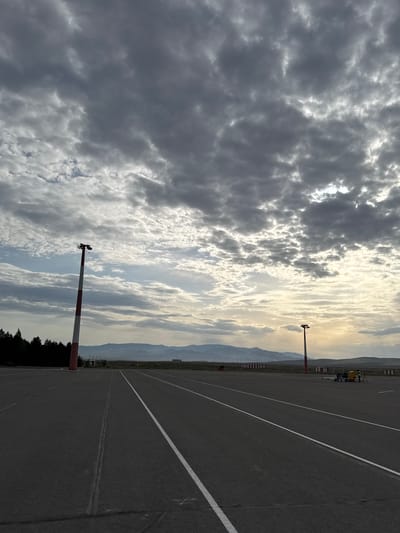 The image depicts an airport runway in Kyzyl, Russia, under a cloudy sky. The runway, made of asphalt, features painted white lines. A red and white striped pole stands along the side of the runway. In the distance, mountains can be seen. The background shows what appears to be airport vehicles and buildings. Trees line the edge of the runway.