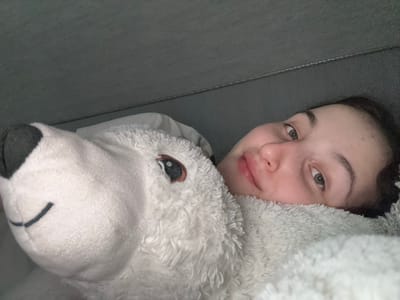 A young woman is in bed with a large stuffed polar bear in Tikhiye Zori, Russia. The woman is lying on her side, with her head resting on the polar bear. Her eyes are open and she is looking at the camera. She is wearing a white, fuzzy blanket. The room is dimly lit.