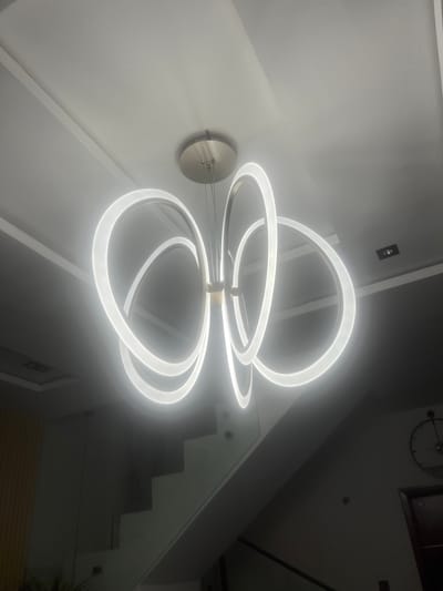 A low-angle indoor shot shows a modern circular chandelier hanging from the ceiling. The chandelier consists of several interconnected illuminated rings. The walls appear to be white or light grey. A ceiling-mounted electrical box and a round, flat ceiling fixture are visible. In the background, a dark staircase ascends along a wall, with a clock and framed picture visible on the upper wall. The environment appears to be residential, though the location of Mopo-Akinlade, Nigeria, cannot be determined from the image alone. There are no people present in the image.