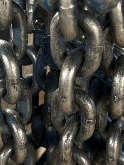 The image displays a close-up, detailed view of a heavy-duty metal chain. The chain links are thick, metallic, and appear to be made of steel, exhibiting signs of wear and tear, with some areas showing a bluish tint, possibly from paint or rust. The lighting highlights the texture and contours of the metal, with reflections and shadows creating a sense of depth. The chains are intertwined and suspended, suggesting they are part of a larger structure or mechanism.
There is no discernible information within the image that indicates a specific location in the city of Lanžhot, Czechia. The scene is purely focused on the chain itself, with no background elements that would provide geographical context. Therefore, it is not possible to determine the location of this image within Lanžhot.