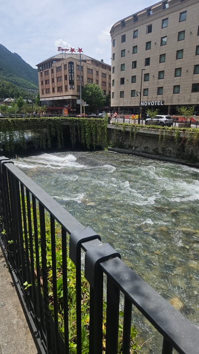 This detailed image depicts a daytime scene along the Gran Valira river in Andorra la Vella, Andorra. In the immediate foreground, a black metal railing with vertical bars and a horizontal top bar extends from the left, positioned above green ground cover and a concrete path.
Beyond the railing, the Gran Valira river flows rapidly from left to right, exhibiting clear water with visible rocks on its bed and several areas of white rapids and turbulence. In the mid-ground, a bridge spans the river, heavily draped with thick green climbing vines that hang downwards towards the water. Above the bridge, a paved street runs parallel to the river, featuring a black metal barrier on its edge, several indistinct pedestrian figures, and parked vehicles, including a white SUV on the right.
The background is dominated by urban structures and natural topography. To the left, a multi-story, light brown building with numerous windows displays a "Bingo" sign topped by two red stars on its roof, and a vertical banner on its facade reading "arthotel" with two "P" symbols below. To the right, a taller, lighter-colored multi-story building with a regular pattern of rectangular windows prominently features a "NOVOTEL" sign on its ground level. In the far background, a steep, densely forested green mountain slope rises on the left side under a bright sky with scattered white clouds.