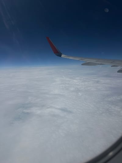 An aerial view from an airplane window shows a vast expanse of white clouds stretching to the horizon, under a deep blue sky. An airplane wing, with a red and white tip, is visible in the foreground, extending from the right side of the frame towards the center. The curvature of the Earth is subtly suggested by the horizon line. There are faint circular reflections in the sky, possibly from the window or a celestial body. The image is devoid of any urban or natural landscape elements that would indicate a specific location within the city of Petrofedorovsk, Russia.