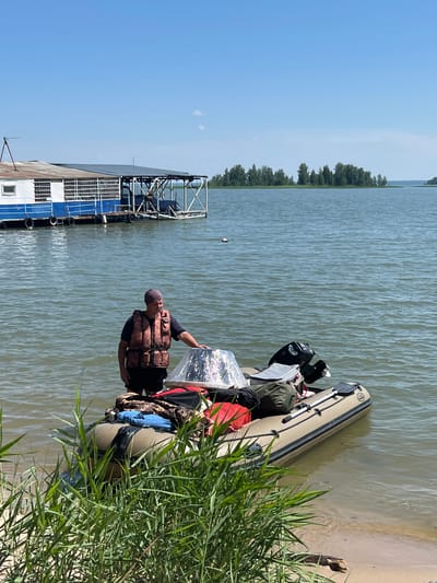 A man wearing a life vest stands in the shallow water next to an inflatable boat loaded with gear, including bags and a cooler. The boat has a motor attached. In the background, a houseboat is docked on a calm body of water under a clear blue sky. The scene is likely in Borovoye, Russia, a resort town known for its natural beauty and lakes.