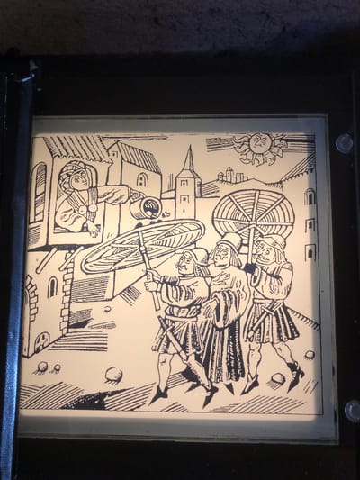 The image depicts an illustration, possibly a woodcut, with a scene reminiscent of medieval art. Three individuals are centrally located, appearing to manipulate large spoked wheels or similar devices. To the left of this central action, architectural elements suggest a town or city, with a person in what seems to be a window or opening in the structure. A stylized sun is positioned in the upper left corner of the image. Scattered throughout the illustration are various geometric shapes.
The image does not contain enough evidence to place it in Veytaux, Switzerland.
