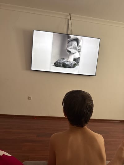 The image shows a young person from behind, watching a television screen mounted on a light-colored wall. The television displays a black and white image of what appears to be a person's feet wearing grey pants and white socks, with a large rock in the foreground. The room has a wooden floor and a white baseboard. A small electrical outlet is visible on the wall below the television. The overall scene suggests a casual indoor setting. The location is identified as Serebryakovka, Russia.