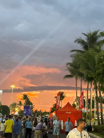 This image depicts a crowd of people gathered outdoors, likely at an event or festival, with palm trees in the background. The sky displays a dramatic sunset with vibrant orange and pink hues, indicating it is either late afternoon or early evening. The lighting is somewhat dim, with some artificial lights from lamp posts visible in the distance.
Several red Coca-Cola branded tents and flags are prominently displayed, suggesting a sponsorship or promotional activity for the beverage company. People are seen standing and mingling, some appearing to be looking in the same direction, possibly at a stage or attraction. The clothing worn by the individuals suggests a warm climate.
The overall atmosphere appears to be festive and casual. Visible text includes "Coca-Cola" on the tents and flags. The location is noted as Miami Gardens, USA, which aligns with the presence of palm trees and the warm weather implied by the attire. The scene captures a moment of gathering and enjoyment during a sunset.
