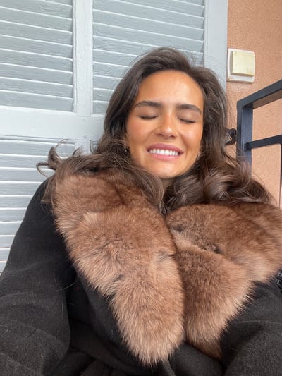 A woman is smiling with her eyes closed. She is wearing a black coat with a large brown fur collar. The woman is positioned in front of a white wooden shutter. The shutter is attached to a building. Behind the shutter there is a pink wall, with a square white plate attached to it. In the background, there is a black metal railing. The image is taken in Roquebrune-Cap-Martin, France. The coordinates are 43.763 latitude and 7.464 longitude.  The photo appears to be taken on a residential property, possibly a balcony.