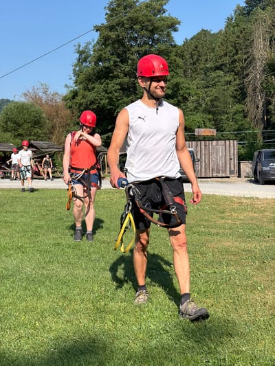 This image depicts an outdoor scene with several individuals dressed in red helmets and harnesses, suggesting participation in an adventure activity such as ziplining or rope climbing. The main focus is on two individuals in the foreground, both wearing red helmets. The person on the right, in the foreground, is a man wearing a white sleeveless shirt, black shorts, and sturdy hiking shoes. He has a harness around his waist with yellow straps attached. The person to his left and slightly behind is a woman wearing a coral-colored tank top, dark shorts, and similar footwear and harness. Both are walking on a grassy area under a clear blue sky, with trees and vegetation in the background. In the middle ground, other individuals can be seen, also equipped with helmets and harnesses, some near wooden structures. The environment appears to be a park or an adventure course, possibly located in the vicinity of Warisy, Belgium, given the context provided. The lighting indicates it is a sunny day, casting shadows on the grass.