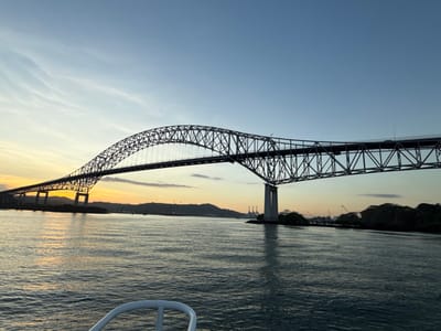 The image captures the Bridge of the Americas, a steel arch truss bridge, spanning the Pacific entrance to the Panama Canal in Ancón, Panama, at sunset. The sky, occupying the upper half of the frame, transitions from deep blue overhead to lighter blue, yellow, and orange hues near the horizon on the left, with faint cirrus clouds present. The bridge is silhouetted in dark tones against the colorful sky, revealing its intricate lattice structure and several vertical support piers extending into the water. Multiple vehicles are discernible as small dark shapes on the bridge's roadway. The lower half of the image displays dark, rippled water, reflecting the sky's ambient light. In the immediate foreground, the white railing structure of a boat is visible at the bottom center, indicating the vantage point. Distant landmasses along both shorelines are visible, featuring silhouetted hills and several industrial cranes on the far banks. No individuals are distinctly identifiable.