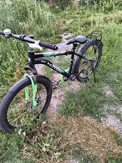 A black YOYODIS MTB mountain bike is parked on a gravel path in a rural area with tall green grass and some dry grass. The bike has green front forks and a black seat. The handlebars are black with white grips and a silver bell. The rear of the bike has a black rack. The bike's frame has a "YOYODIS" logo in white with teal accents. The front wheel has colorful spokes. The background shows more grass and foliage, suggesting an outdoor, natural environment. There are no people visible in the image. The location is described as Mayma, Russia, consistent with a rural setting.