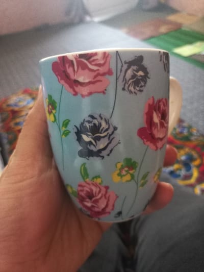 A human right hand, with a light skin tone, holds a light blue ceramic mug from the left side. The mug is decorated with a floral pattern featuring large pink and dark grey roses, interspersed with smaller yellow and green flowers and green stems. A segment of the mug's white handle is visible on the right. In the blurred background, a section of dark grey or blue denim fabric, consistent with clothing, is present in the lower right. To the left and behind the hand, a multicolored textile with distinct red, yellow, and blue patterns is discernible. The scene is an indoor setting in Navbahor, Uzbekistan, with indistinct soft furnishings in the background and a light source originating from the upper right.