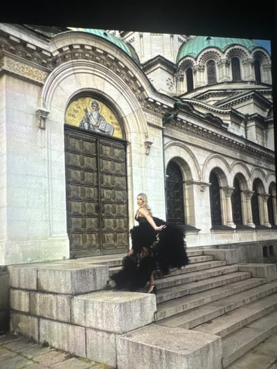 The image depicts the Alexander Nevsky Cathedral in Sofia, Bulgaria. A woman in a large, dark-colored dress stands on the steps leading to one of the building's entrances. The cathedral's architecture, including its arches, domes, mosaic above the doorway, and the steps are visible.