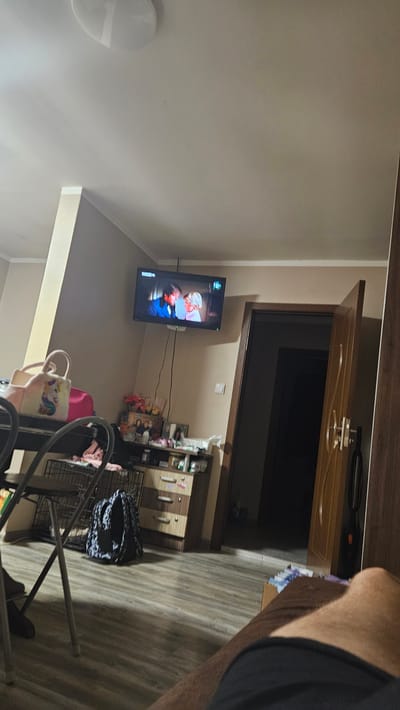 The image presents an indoor domestic scene, likely a living room or common area, captured from a low, tilted perspective. The room features light-colored, possibly beige or off-white, walls and a light wood-patterned laminate floor.
A flat-screen television is mounted on the left wall, displaying a colorful animated program or cartoon with two prominent characters, one dressed in red and white, and another in blue. Black electrical cables are visible running from the TV. To the right of the television, a shelving unit or cabinet holds various items, and below it, a dark wire crate, possibly a pet carrier, is present. In the foreground, near the bottom center, a pink tote bag with a unicorn design and another light-colored bag are visible.
In the background, a wooden door frame and a partially open wooden door lead into what appears to be a darker room. A round, white ceiling light fixture is visible on the upper left side of the frame. On the far right, a person's bare leg and part of an arm wearing a dark shirt are visible, indicating the photograph was taken from their perspective, likely while they were seated or resting.
The scene suggests an activity of watching television in a relaxed home environment. The internal lighting conditions, with no visible external light source, imply it could be evening or night. The setting is a typical home interior, consistent with the location context of Slivo Pole, Bulgaria. No discernible text is visible anywhere in the image.