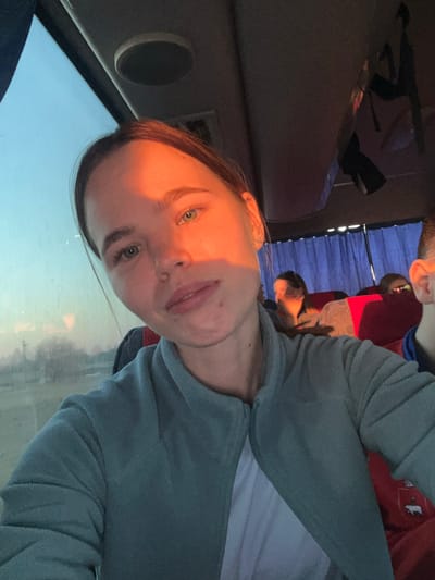 A selfie photograph taken inside a vehicle. The main subject is a person with fair skin and light-colored eyes, wearing a gray-blue long-sleeved shirt over a white t-shirt. The person's face is illuminated by sunlight, creating shadows. Other people are visible in the background, sitting behind blue window coverings. The vehicle appears to be in motion, as suggested by the blurry view through the window. The location, Ustinovo, Russia, cannot be confirmed.