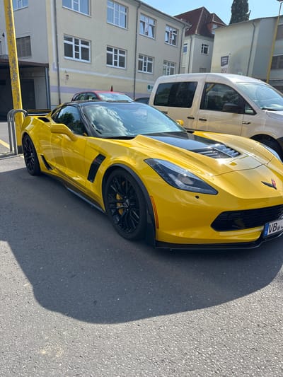 The image shows a bright yellow Chevrolet Corvette parked on an asphalt road in front of a light-colored building. The car is the primary subject, and it's viewed from a low angle, emphasizing its sporty design. The car has black rims and a black grille. A license plate is visible on the front of the car, with the text "VB" and other characters. To the left of the Corvette, a white van is partially visible. The building in the background has multiple windows and appears to be a residential or commercial building. The scene is lit by bright sunlight, casting a long shadow from the car onto the road, suggesting it's daytime. The weather appears to be clear and sunny. There are no people visible in the image. The location context is Alsfeld, Germany.
