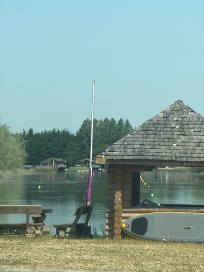 The image shows a boathouse and pier on a lake in the Cotswold District, United Kingdom. The boathouse has a grey shingled roof and is situated on the edge of the water. A silver-colored pole with a purple cloth attached is visible near the building. A grey paddleboard rests on the grassy bank next to the boathouse. In the background, trees are visible. The lake reflects the boathouse, trees, and sky. The foreground features a pier with wooden supports and logs.