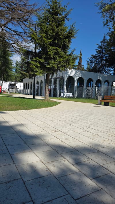 The image depicts a paved walkway in front of a modern building with arched windows and entrances, located in Babyshira, Abkhazia. The building appears to be a public space or commercial establishment. Several evergreen trees are scattered around the scene, with one prominent tree standing near the walkway and building. The sky is clear and blue, with a visible moon. A wooden bench and a trash receptacle are situated on the right side of the walkway, indicating a public park or recreational area. The ground is covered with light-colored rectangular paving stones, and shadows from the trees are cast across them, suggesting it is a sunny day. The overall atmosphere is calm and serene.